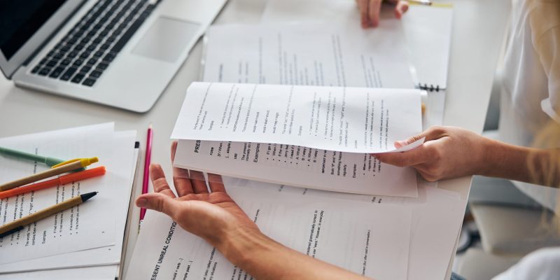 Parent helping her teenage daughter with understanding the grammatical topic from a schoolbook while working at a cluttered desk